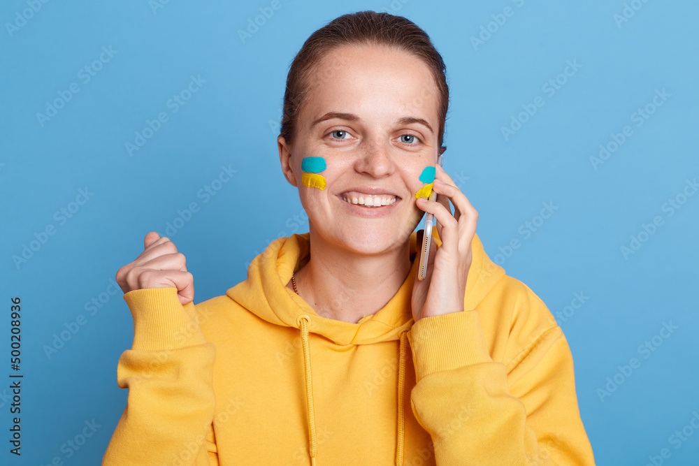 Delighted smiling woman in yellow hoodie with Ukrainian flag on cheeks, posing isolated over blue background, talking on cell phone, clenched fists, hearing great news. Delighted smiling woman in yellow hoodie with Ukrainian flag on cheeks, posing isolated over blue background, talking on cell phone, clenched fists, hearing great news.