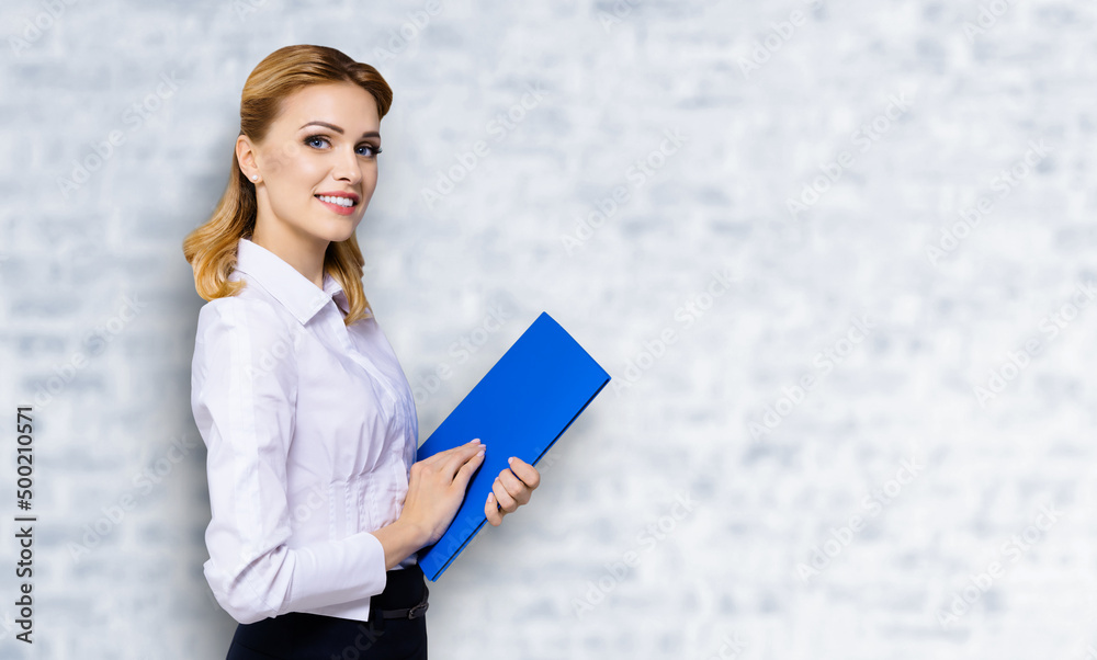 Happy smiling attractive businesswoman, with copy space area for some slogan or text. Confident blond woman in white shirt with blue folder, over white bricks wall. Business concept. Happy smiling attractive businesswoman, with copy space area for some slogan or text. Confident blond woman in white shirt with blue folder, over white bricks wall. Business concept.