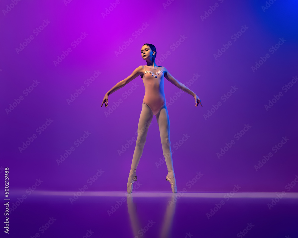 Studio shot of beautiful school age girl, ballet dancer dancing isolated on purple background in neon light. Art, grace, beauty, ballet school concept