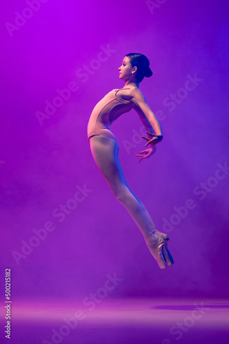 Portrait of young little ballet dancer, teen jumping isolated on purple background in neon light. Art, grace, beauty, ballet school concept © master1305