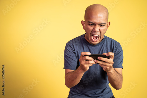 Bald asian man shouting with mouth open while playing game using smartphone on isolated background © Odua Images