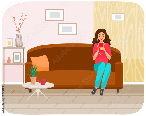 Woman sitting on sofa holds smartphone in her hand. Casual lady in headpones listening to music and browsing social media on mobile device. Girl on couch uses phone for chatting and surfing internet