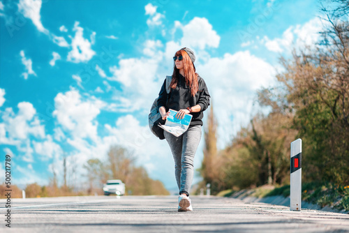 A young cacuasian hipster woman walks along the road with a map and backpack in her hands. Copy space. Concept of hitchhiking and local travel © _KUBE_