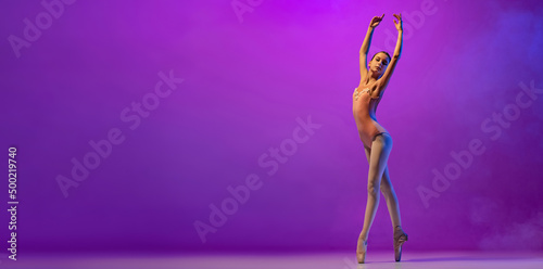 Beautiful flexible young female ballet dancer, teen in stage outfit and pointes dancing isolated on purple background in neon light with smoke. Art, grace, beauty. Flyer
