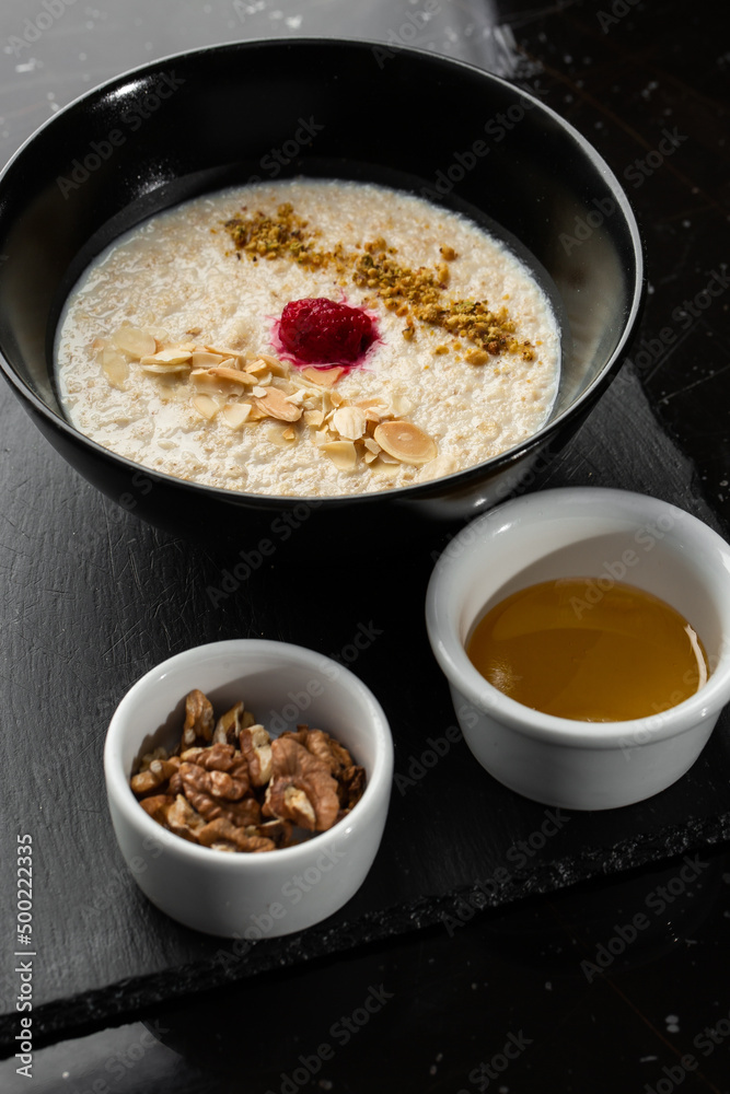 Semolina porridge for breakfast oat with nuts in black bowl served with honey isolated on black stone background. Homemade food. Tasty breakfast. Semolina porridge for breakfast oat with nuts in black bowl served with honey isolated on black stone background. Homemade food. Tasty breakfast.