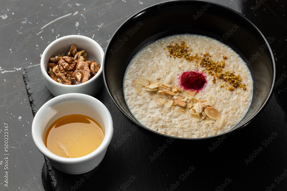 Semolina porridge for breakfast oat with nuts in black bowl served with honey isolated on black stone background. Homemade food. Tasty breakfast. Semolina porridge for breakfast oat with nuts in black bowl served with honey isolated on black stone background. Homemade food. Tasty breakfast.