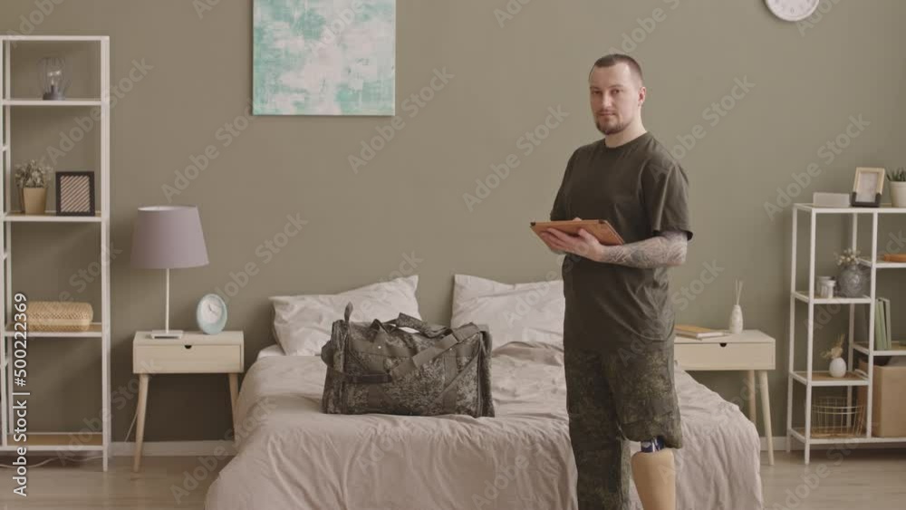 Medium slowmo of young adult Caucasian man with prosthetic leg in military uniform using tablet pc then looking at camera standing in his bedroom with olive green walls and army bag on bed Medium slowmo of young adult Caucasian man with prosthetic leg in military uniform using tablet pc then looking at camera standing in his bedroom with olive green walls and army bag on bed