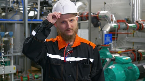 Industrial engineer at a manufacturing plant. A bearded man in a white hard hat and overalls at the workplace. Work in the oil and gas structure at the oil and gas processing plant. © ShantiMedia