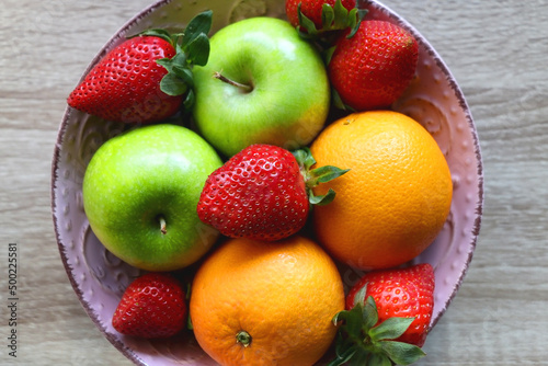 Pink bowl filled with fresh apples  oranges and strawberries on wooden table. Flat lay.