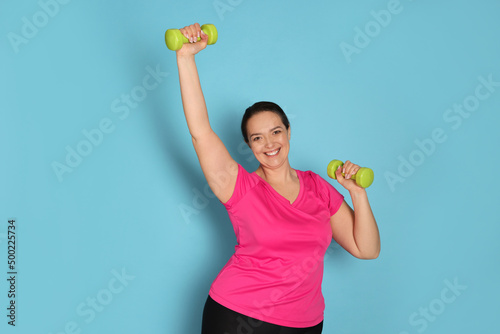 Happy overweight woman doing exercise with dumbbells on light blue background