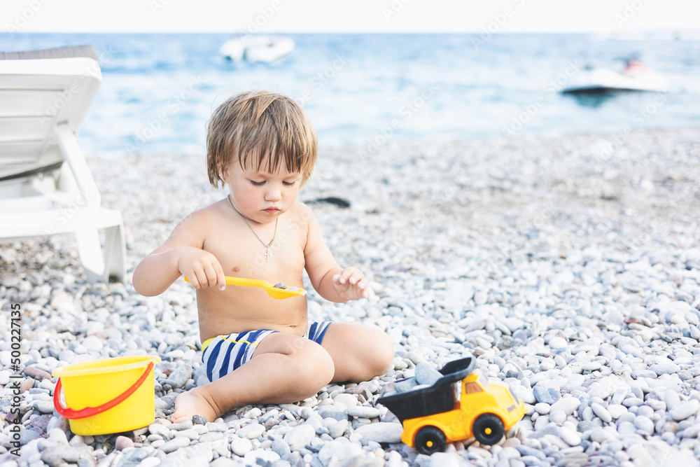 Cute child playing with yellow bucket and toy car on pebble beach. Happy toddler on sea coast. Summer family holiday vacation. Two years old little boy having fun. Summertime. Travel concept Cute child playing with yellow bucket and toy car on pebble beach. Happy toddler on sea coast. Summer family holiday vacation. Two years old little boy having fun. Summertime. Travel concept