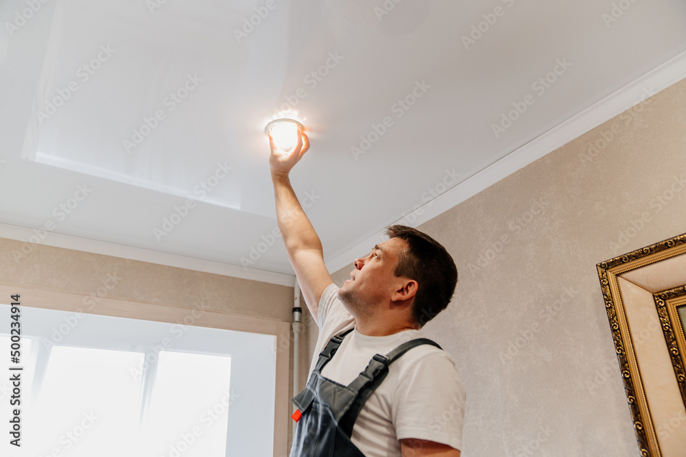 Fototapeta premium A male electrician changes the light bulbs in the spotlight ceiling lamp
