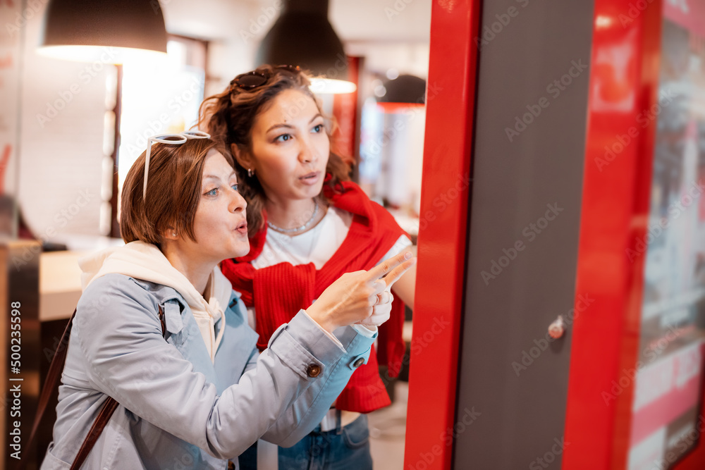 Two girl friends orders food and lunch at a fast food restaurant using a self-service kiosk or a terminal with a screen. Modern commerce equipment