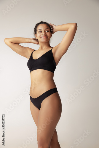 Portrait of young woman with natural body posing in black underwear isolated over grey studio background. Femininity