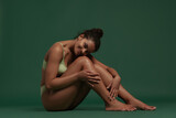 Portrait of young tender woman sittin on floor, posing in underwear isolated over green background. Anti-cellulite care