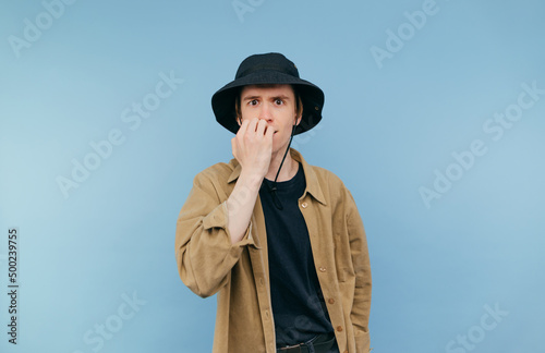Nervous guy in panama and shirt looks at the camera and bites his nails from unpleasant experiences on a blue background.