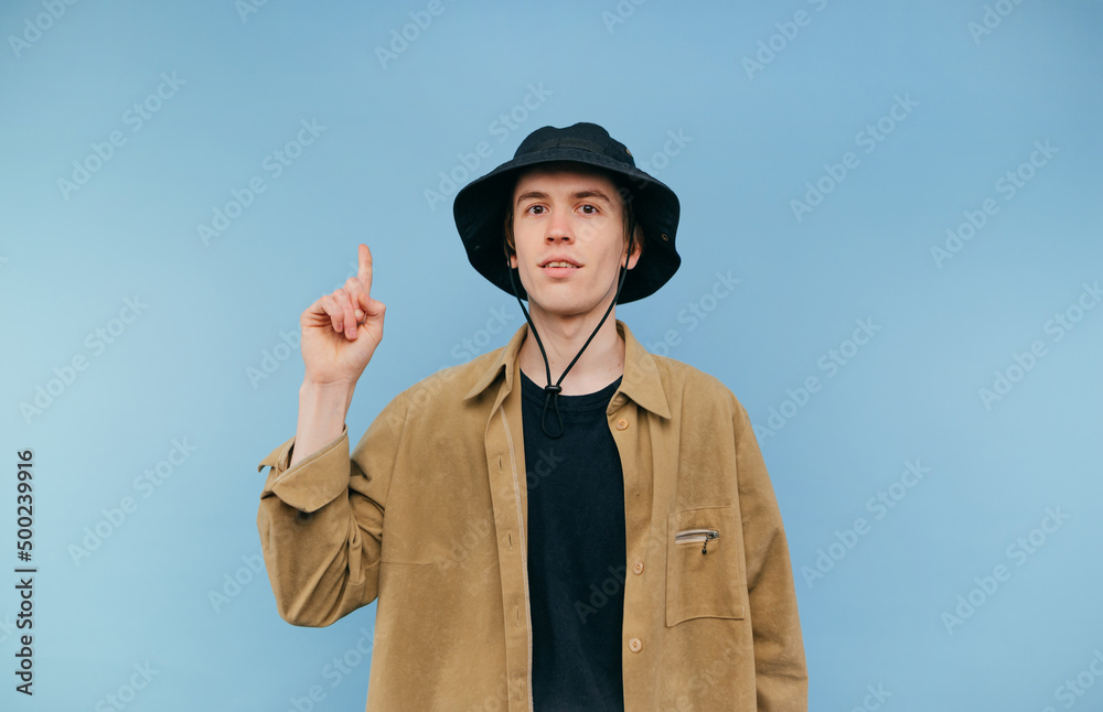 Positive hipster in a shirt and panama stands on a blue background and points his finger up at an empty spot with a smile on his face. Positive hipster in a shirt and panama stands on a blue background and points his finger up at an empty spot with a smile on his face.