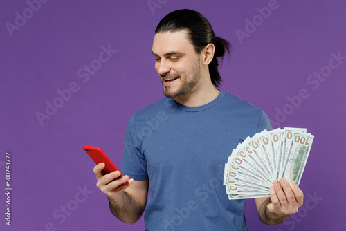 Young smiling man 20s wear basic blue t-shirt hold mobile cell phone holding fan of cash money in dollar banknotes isolated on plain purple color background studio portrait. People lifestyle concept.