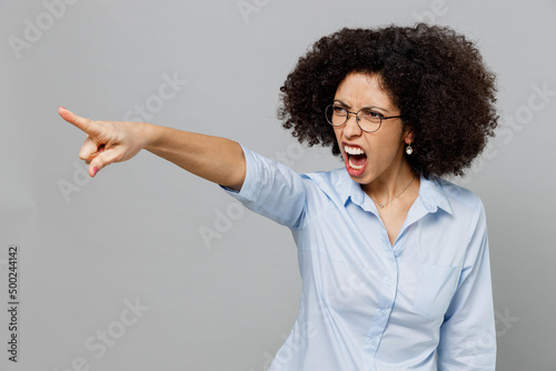 Young angry sad employee business corporate lawyer woman of African American ethnicity in classic formal shirt work in office point index finger aside scream isolated on grey color background studio.