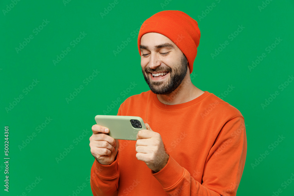 Young gambling happy cool man 20s wear orange sweatshirt hat using play racing app on mobile cell phone hold gadget smartphone for pc video games isolated on plain green background studio portrait Young gambling happy cool man 20s wear orange sweatshirt hat using play racing app on mobile cell phone hold gadget smartphone for pc video games isolated on plain green background studio portrait