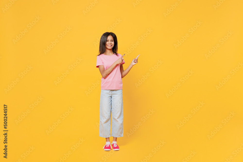 Full body fun little kid girl of African American ethnicity 12-13 years old in pink t-shirt point index finger adide on workspace area isolated on plain yellow background. Childhood lifestyle concept. Full body fun little kid girl of African American ethnicity 12-13 years old in pink t-shirt point index finger adide on workspace area isolated on plain yellow background. Childhood lifestyle concept.