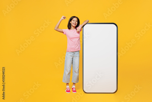 Full body little kid girl of African American ethnicity 12-13 years old in pink t-shirt big huge blank screen mobile cell phone workspace area do winner gesture isolated on plain yellow background