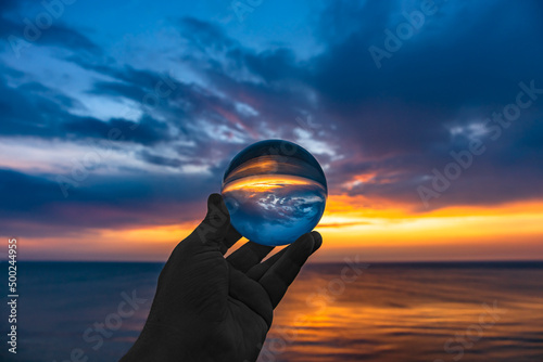Beautiful scenic view through lens of crystal ball on water horizon at summer sunset. Clouds reflection in sphere. Crystal ball in hand holding globe in outdoor.Blurred background.