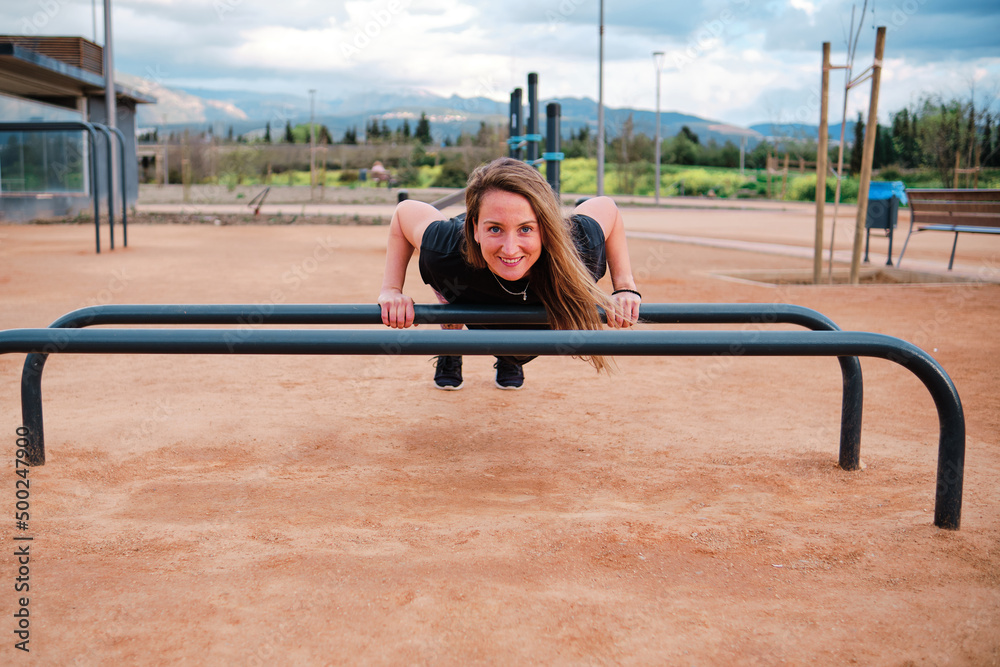 Young blonde woman performing calisthenics exercises in an urban park. She is exercising on the parallel bars