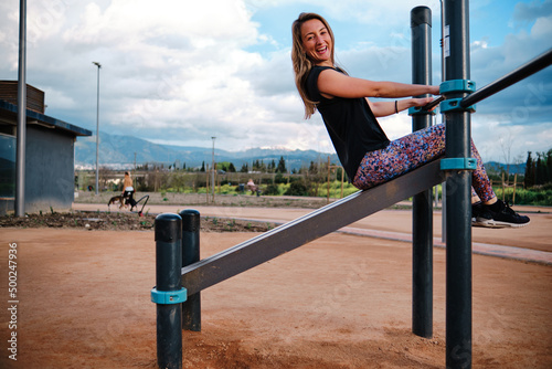 Young blonde woman performing calisthenics exercises in an urban park. She is doing sit-ups on the bench