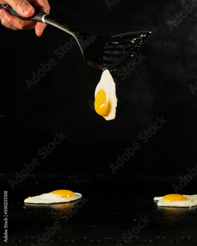 three eggs sunny side up with two on the griddle and one being flipped on a black background