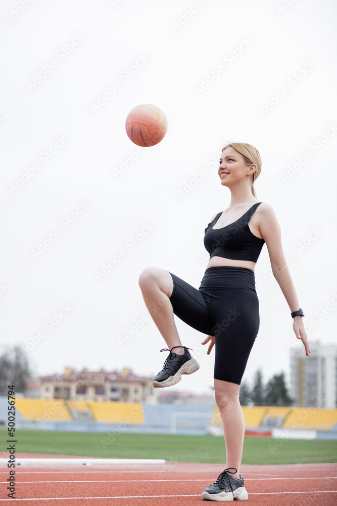 full length view of happy woman in black sports bra and bike shorts training with ball.
