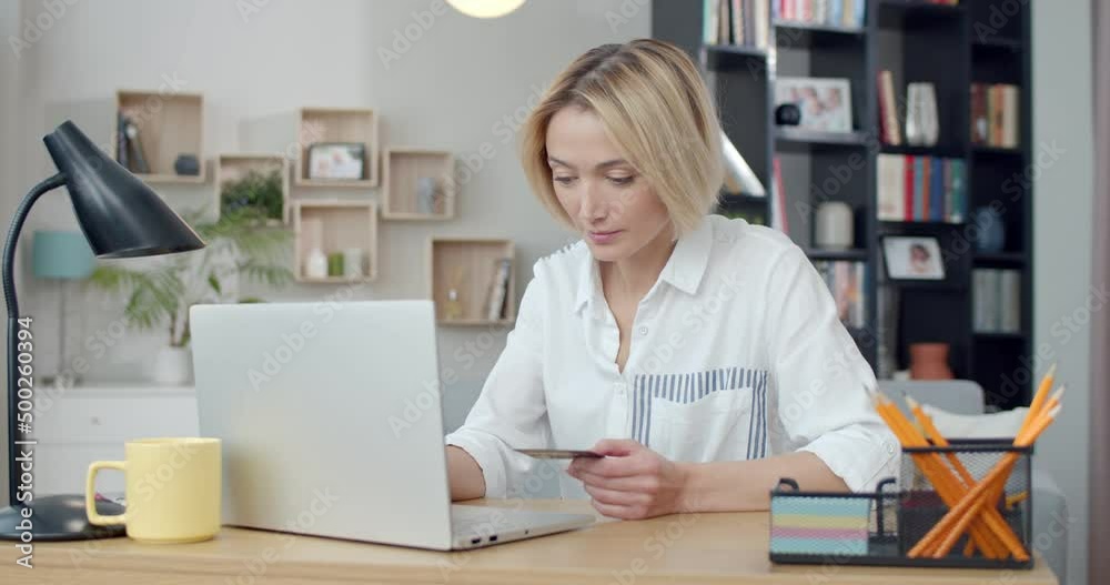 Positive young woman using modern laptop and credit card for online shopping at home. Happy caucasian blonde sitting at desk and making purchases in internet. Positive young woman using modern laptop and credit card for online shopping at home. Happy caucasian blonde sitting at desk and making purchases in internet.