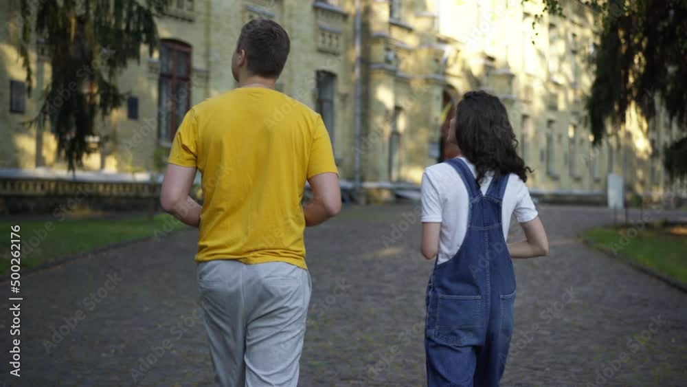 Tracking shot of Caucasian groupmates talking walking at university campus in the morning. Back view young confident smart man and woman strolling outdoors discussing studies. Intelligence concept Tracking shot of Caucasian groupmates talking walking at university campus in the morning. Back view young confident smart man and woman strolling outdoors discussing studies. Intelligence concept