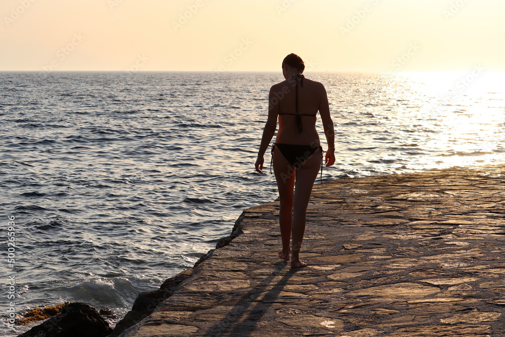 Silhouette of lonely woman in bikini walking by the stone beach on sunset and sea background. Romantic vacation and swimming concept