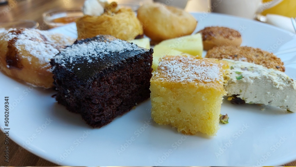 Dessert auf einem weißen Porzellanteller. Orientalische Kekse. Schokoladenwürfel mit Kokos. Kadaif mit Walnüssen. Donuts. Sesamkuchen. Gläser mit Fruchtmarmelade. Kanafeh-Kuchen.