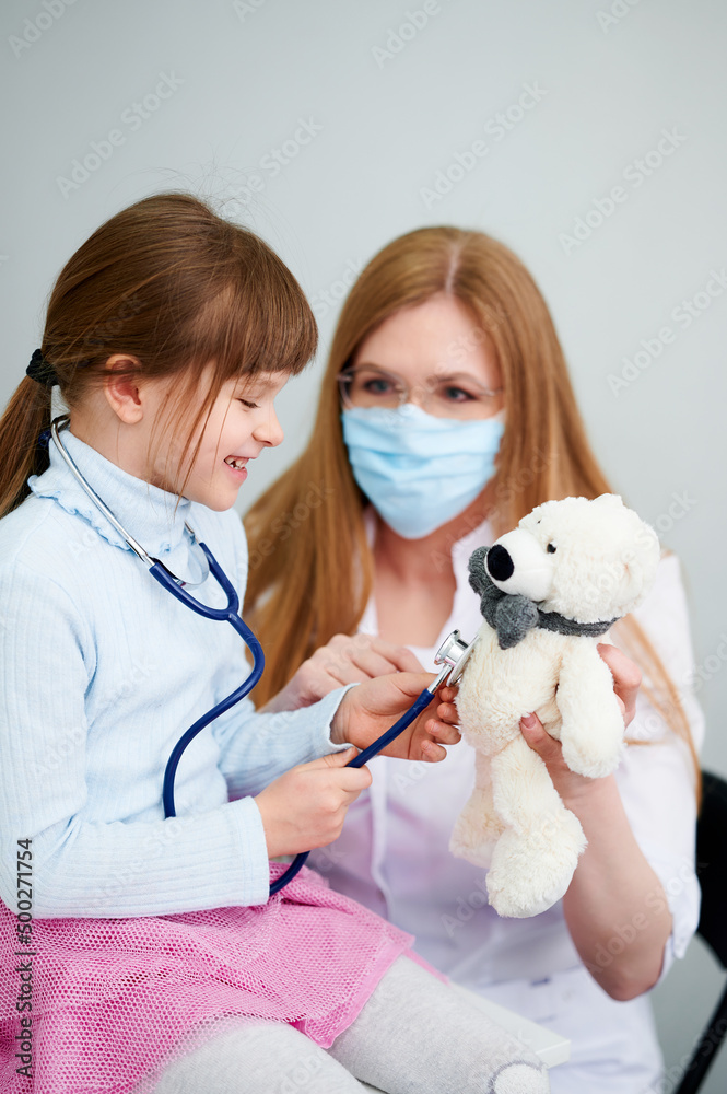 Caucasian female doctor wear face mask show little girl examining bear toy by stethoscope indoor hospital Caucasian female doctor wear face mask show little girl examining bear toy by stethoscope indoor hospital