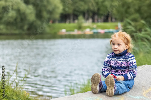 A little girl toddler is sitting on the shore of a lake in the spring forest. Nature walk