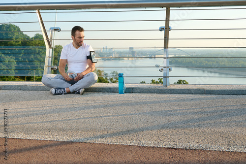 Middle aged Caucasian man in sportswear sitting on a city bridge and checks a mobile application with monitoring heart rate and calories burned during a workout on his phone in a smartphone holder