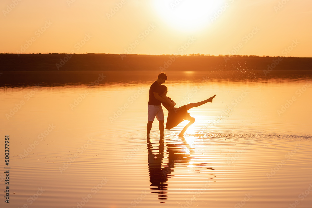 Silhouette of a couple in love, dances with splashes in the water of the lake in the orange sunset