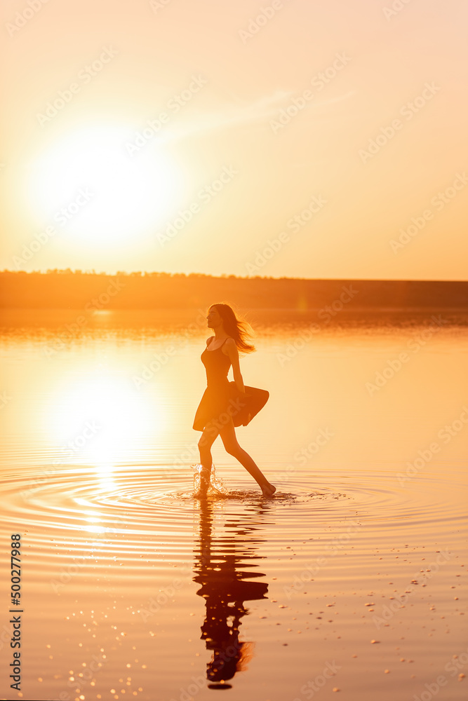 Silhouette of a young woman in an airy black dress in the water of a lake with sky reflection. Silhouette of a young woman in an airy black dress in the water of a lake with sky reflection.