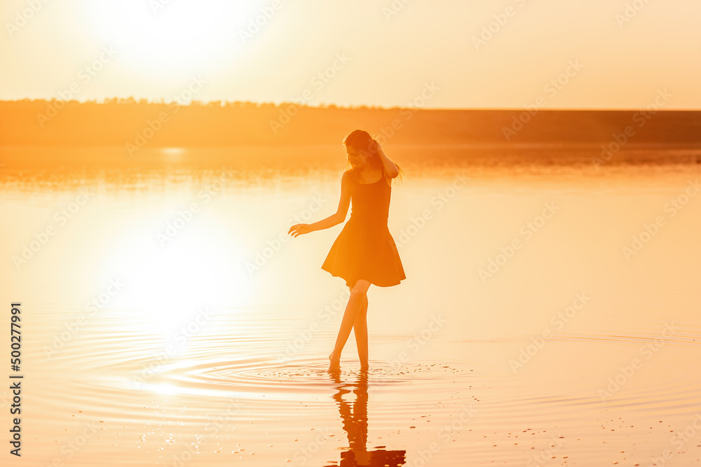 Silhouette of a young woman in an airy black dress in the water of a lake with sky reflection.