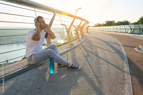 Attractive relaxed middle aged muscular man putting on headphones and listening to music, sitting on a bridge and enjoying the rest after morning workout. Beautiful sun rays falling on the treadmill