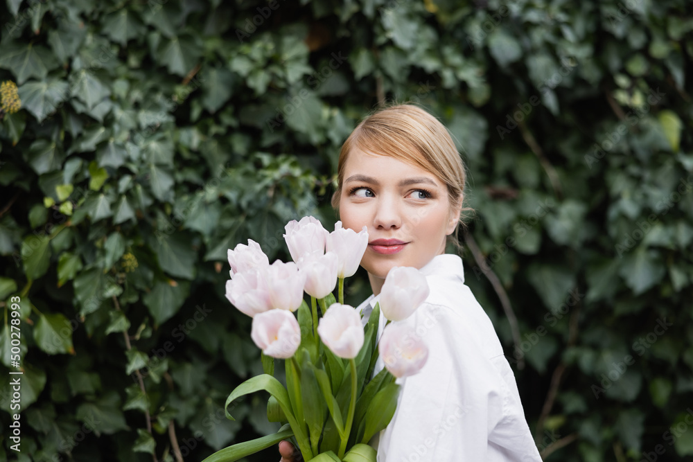 young blonde woman with white tulips smiling near green ivy outdoors. young blonde woman with white tulips smiling near green ivy outdoors.