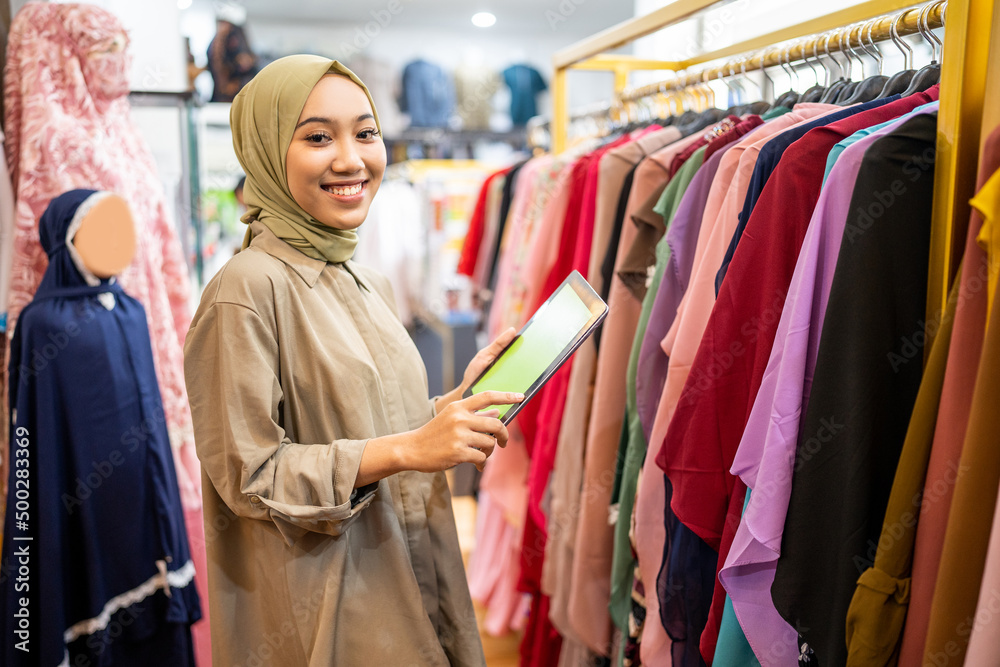 business owner wearing hiijab using a tablet while holding clothes on a hanger at a Muslim clothing store business owner wearing hiijab using a tablet while holding clothes on a hanger at a Muslim clothing store
