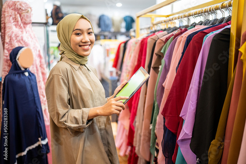 business owner wearing hiijab using a tablet while holding clothes on a hanger at a Muslim clothing store