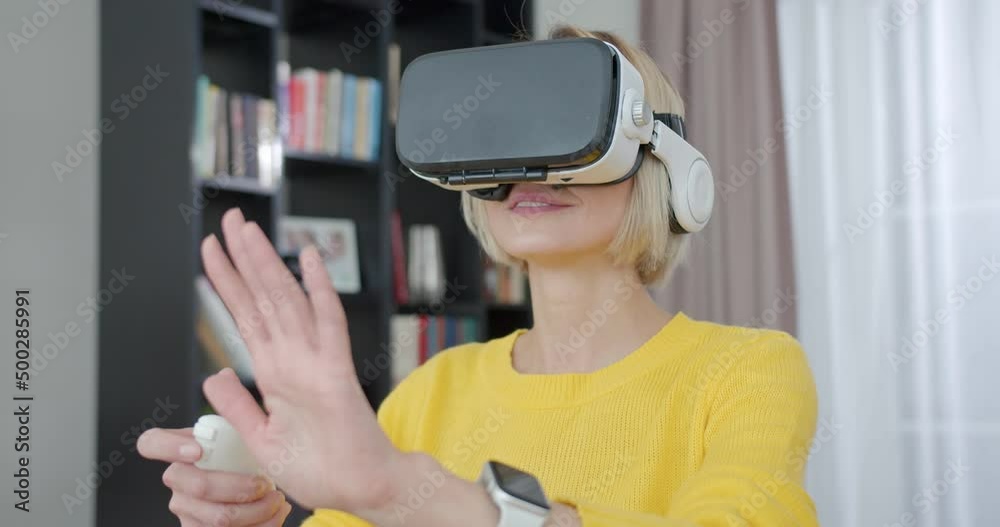 Excited young lady with blond hair wearing virtual reality headset and holding remote controller during leisure time at modern apartment. Concept of people, entertainment and technology. Excited young lady with blond hair wearing virtual reality headset and holding remote controller during leisure time at modern apartment. Concept of people, entertainment and technology.