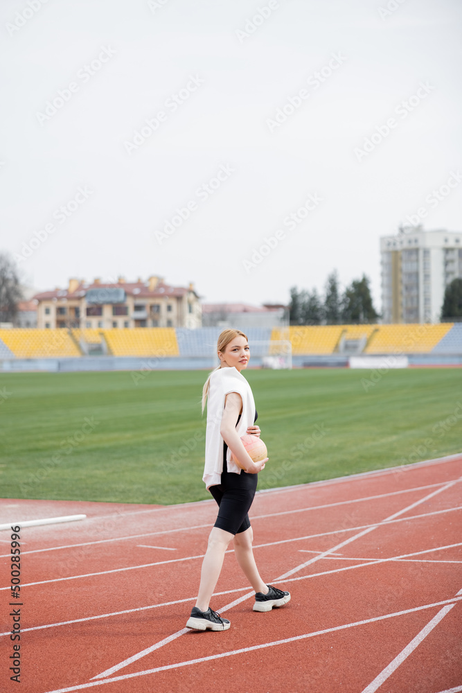 full length view of young sportswoman looking at camera while walking with ball on stadium.