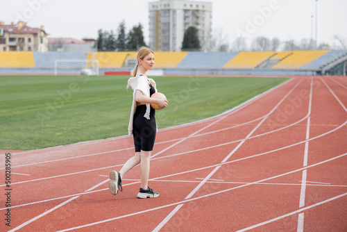 full length view of sportive woman walking on athletic field with ball.