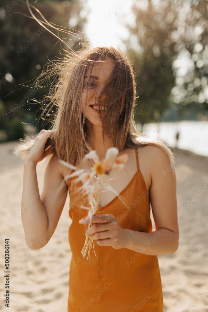 Lovely romantic girl with dark hairstyle dressed orange summer dress is looking at camera with flying hair and holding cottonweed Lovely romantic girl with dark hairstyle dressed orange summer dress is looking at camera with flying hair and holding cottonweed