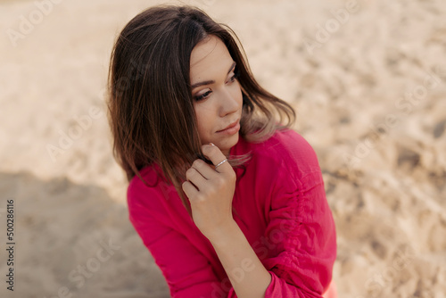 Top view photo of romantic pretty girl with dark hair is looking aside and touching chin while resting outdoor in sunshine on sandy beach